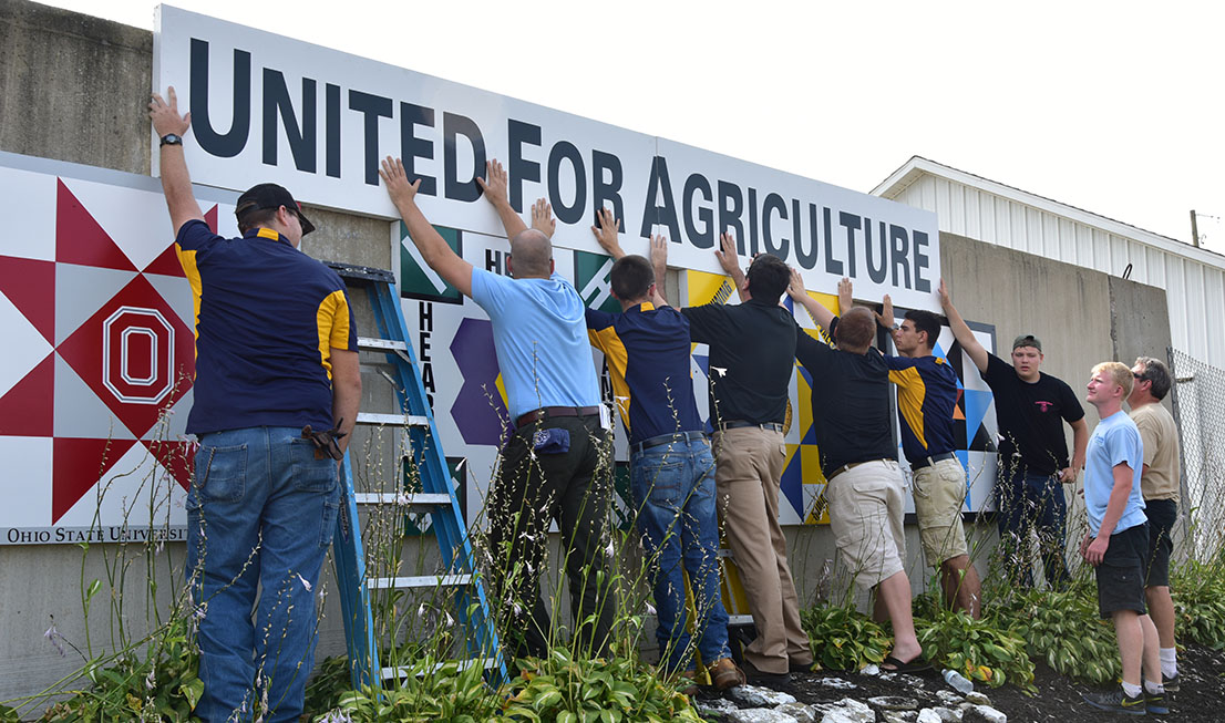 Volunteers and students work to get the banner symmetrically hung over the barn quilts.